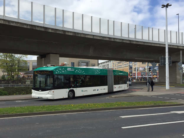 Ein grüner und weißer Bus fährt unter einer Brücke auf einer Straße in der Stadt, mit Menschen auf dem Gehweg, Laternenmästen, Ampeln und Gebäuden mit Fenstern im Hintergrund, unter einem bewölktem Himmel und neben einer Straßenbegrenzung.