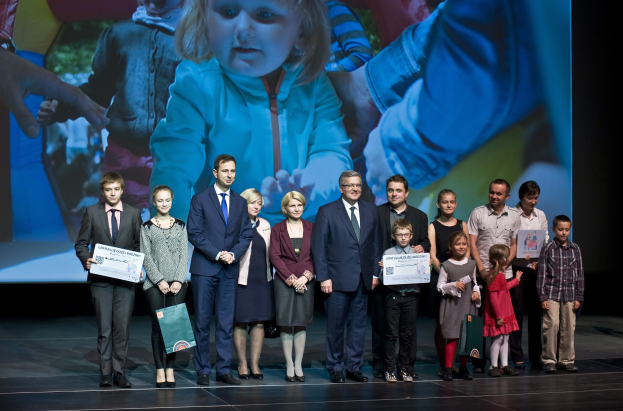 Gruppe von Menschen auf einer Bühne mit Taschen und Urkunden, im Hintergrund ein Bildschirm mit dem Bild eines Kindes und anderen Personen.