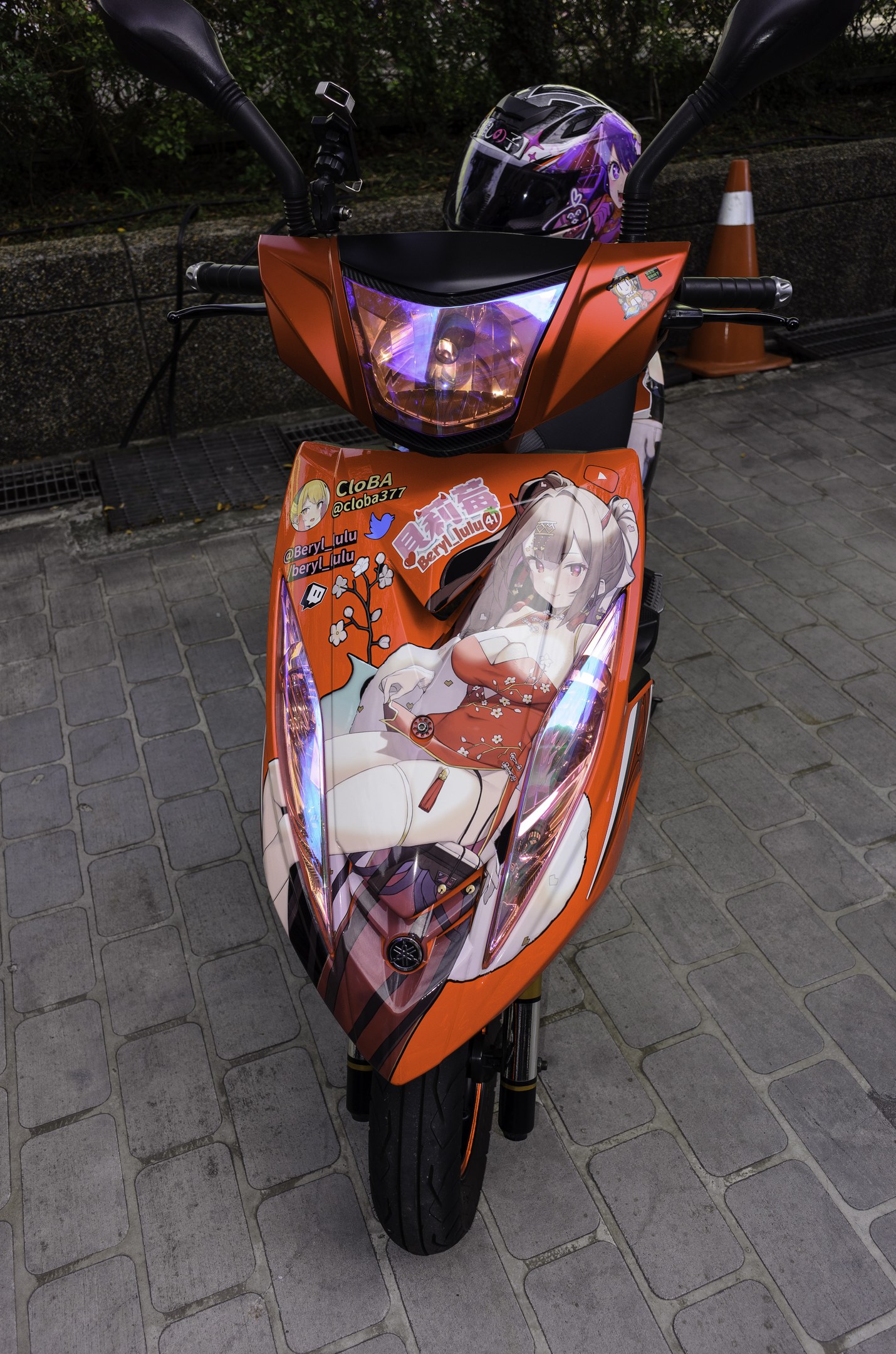 Orangeroller mit Helm auf der Straße geparkt, neben einer Verkehrskegel, einer Wand und Bäumen im Hintergrund.