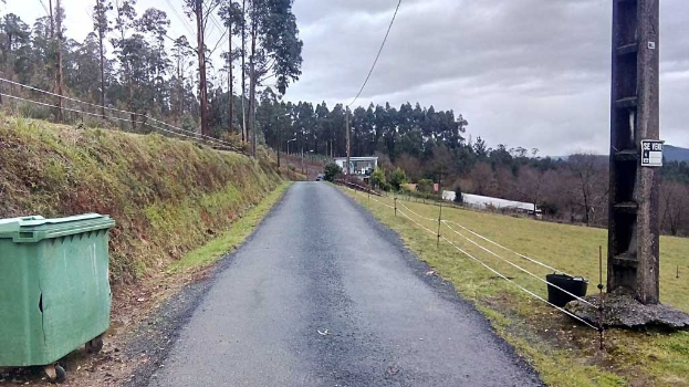 Grüner Müllcontainer an der Seite einer Straße mit Gras, Pflanzen, Bäumen, Masten, Drähten und einem Zaun, unter einem bewölkten Himmel, mit einer mastmontierten Tafel auf der rechten Seite und verstreuten Gegenständen auf der linken Seite.