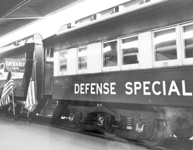 Schwarz-weiß-Foto eines Zuges mit der Aufschrift "Defense Special" auf den Schienen mit zwei Fahnen links und einer Decke oben.