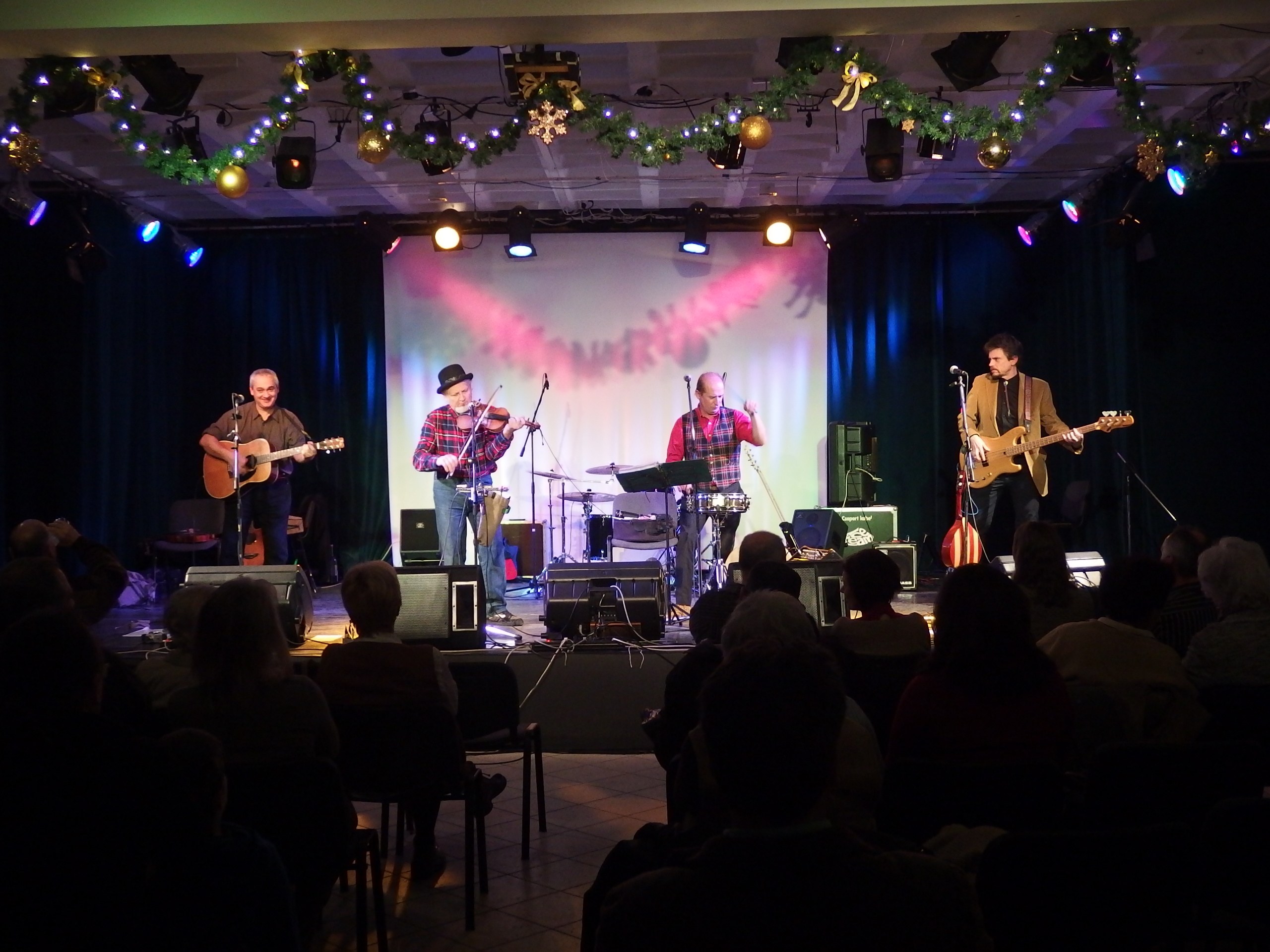 Eine Gruppe von Menschen, die auf der Bühne mit Musikinstrumenten, Mikrofonen und Ständern spielt, während das Publikum auf Stühlen sitzt; Bühnenlichter und dekorative Elemente sind oben sichtbar.