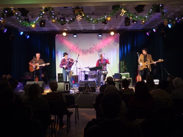 Eine Gruppe von Menschen, die auf der Bühne mit Musikinstrumenten, Mikrofonen und Ständern spielt, während das Publikum auf Stühlen sitzt; Bühnenlichter und dekorative Elemente sind oben sichtbar.