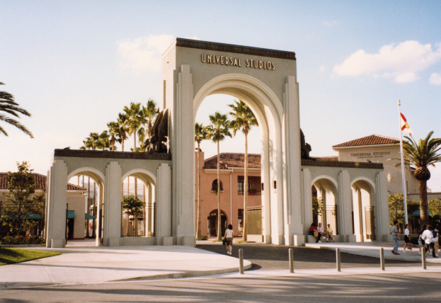 Eingang zu Universal Studios Hollywood mit einem Bogen mit Text, einer Statue, Menschen, Pfählen, Grün, Gebäuden, einem Fahnenmast mit einer Fahne.