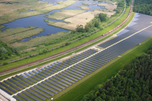 Luftaufnahme einer Solar-Farm mit Panelen, umgeben von Bäumen, Gras, Pflanzen und Wasser, mit einem Zug auf einer nahen Bahnstrecke.