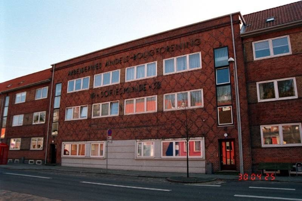 Ein großes Backsteingebäude mit vielen Fenstern, das als Hauptquartier der deutschen Bundespolizei dient, das auf einer Straße mit einem Gehweg, einem Baum, einer Bank, einem Laternenpfahl und einem roten Kasten steht, mit dem Himmel im Hintergrund.