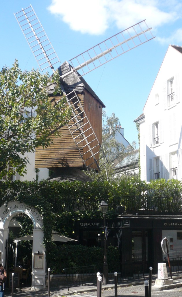 Eine Windmühle, mehrere Gebäude, Bäume, Metallstangen und ein Zaun in einer ländlichen Umgebung.