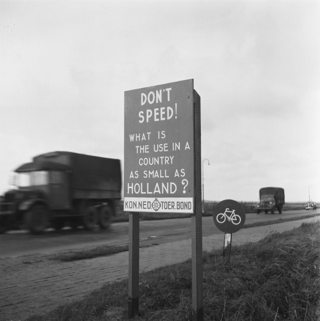 Schwarz-weißes Foto eines Lkw, der auf einer Straße mit einem "Fahren Sie nicht zu schnell"-Schild im Vordergrund und anderen Fahrzeugen, Gras, Pfosten und einem klaren Himmel im Hintergrund fährt.