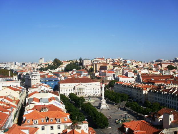 Blick auf Lissabon von einem Hügel mit Gebäuden, Bäumen, einer Statue auf einem Sockel, einer Straße mit Menschen und dem Himmel im Hintergrund.