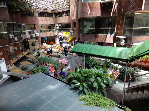 Ein geräumiger Atrium mit viel Grün, Menschen, Tischen, Stühlen und Treppen mit Geländern, vor Gebäuden mit Fenstern, Klimaanlagen und einem Schuppen im Hintergrund.