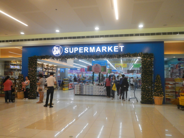 Eine Gruppe von Menschen vor einem Supermarkt, einige mit Einkaufstägern, einem Einkaufswagen, einer Topfpflanze, Regalen mit Gegenständen, Schildern und einer Decke mit Leuchten.