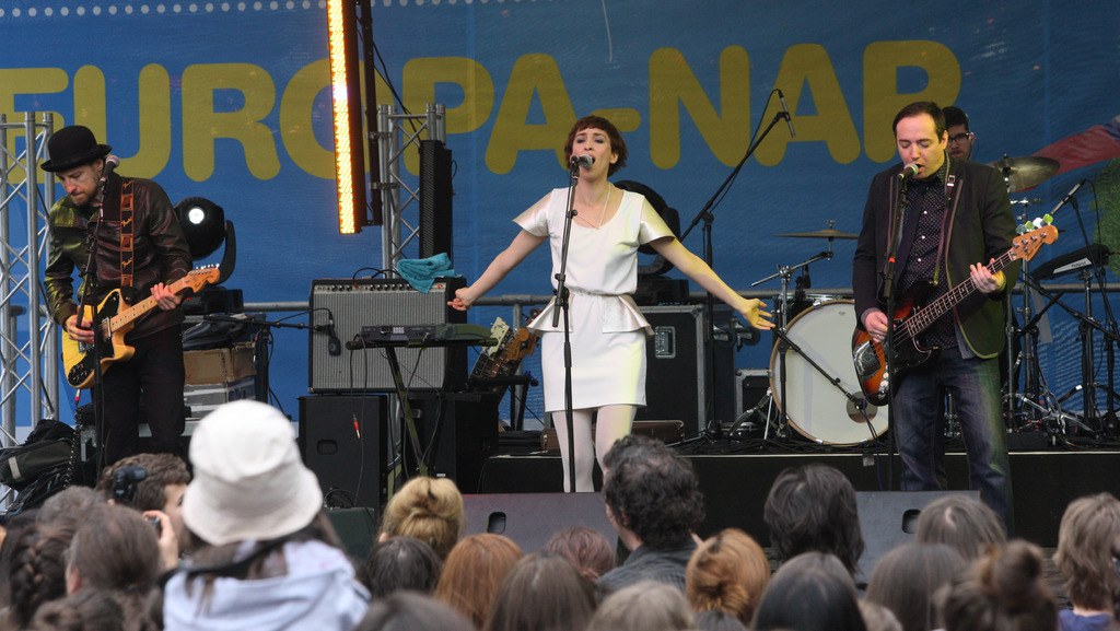 Vier Personen auf der Bühne: Eine Frau in einem weißen Kleid singt in der Mitte, während zwei Männer links und rechts Gitarren spielen; eine Menge ist unten sichtbar, mit Lautsprechern und einer Wand im Hintergrund.