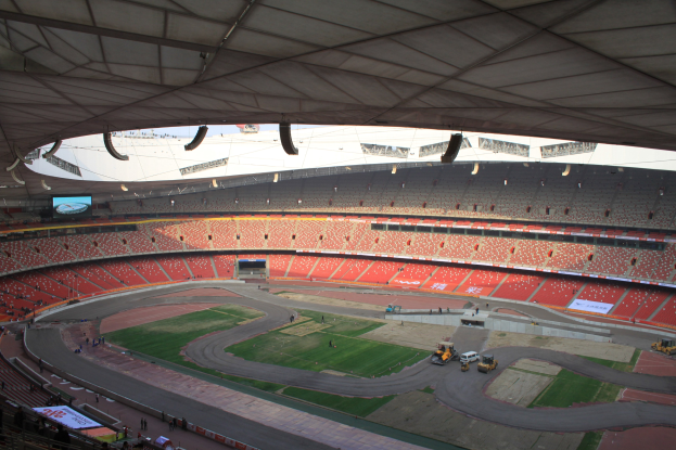 Großes Stadion voller Zuschauer, Fahrzeuge auf der Straße, grasbewachsener Platz, Texttafeln, Geländer und Deckenbeleuchtung.