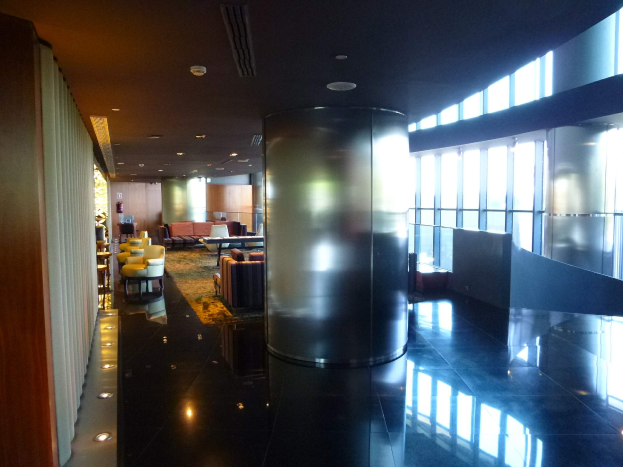 Eine geräumige First-Class-Lounge am Hong Kong International Airport mit großen Fenstern, hohen Decken mit elektrischen Leuchten, Sitzanordnungen einschließlich Stühlen und Sofas, Tischen, einem Teppichboden und einer zentralen Säule.