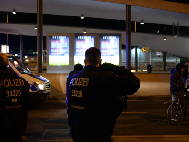 Eine Gruppe von Polizisten vor einem Gebäude in der Nacht, mit einem geparkten Auto links und einem Radfahrer rechts, beleuchtet von Bildschirmen und Lichtern im Hintergrund.