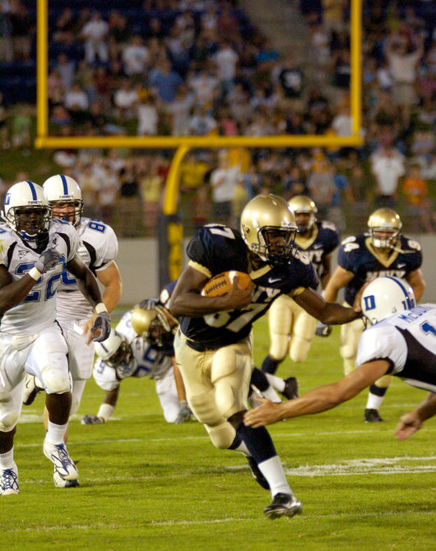 Ein Füßballspieler in einem Sportdress und Helm rennt mit dem Ball, umgeben von anderen Spielern, mit unscharfem Hintergrund und Text unten im Bild.