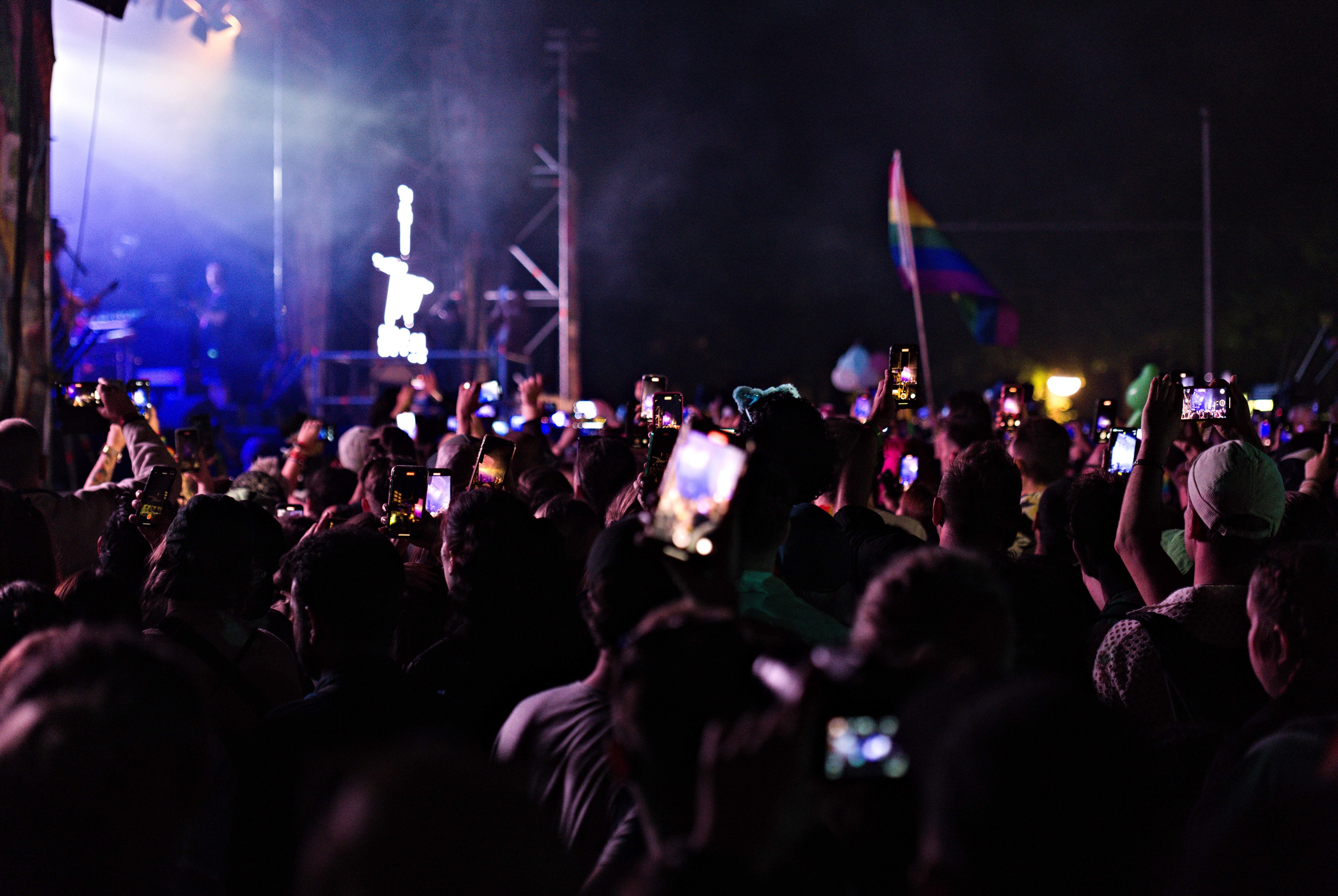 Eine Menge bei einem Konzert mit hochgehaltenen Handys, beleuchtet von Lichtern, umgeben von Pfählen und einer Flagge, während Musiker auf der Bühne spielen.