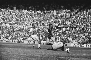 Schwarß-weißes Foto von Männern, die auf einem Feld Füssball spielen, mit Zuschauern im Hintergrund und einer "Bundesliga 1978-79 Ajax 1-1 Hannover 96"-Tafel.