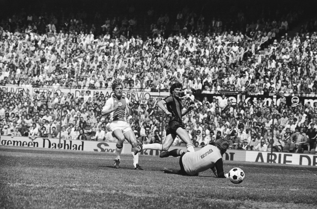 Schwarß-weißes Foto von Männern, die auf einem Feld Füssball spielen, mit Zuschauern im Hintergrund und einer "Bundesliga 1978-79 Ajax 1-1 Hannover 96"-Tafel.