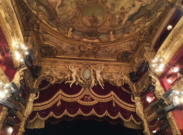 Theaterinnenraum mit roter Gardine, Kronleuchtern, Deckenplastiken, Lampen und einem Gemälde, das eine Bühne und einige Personen unten zeigt.