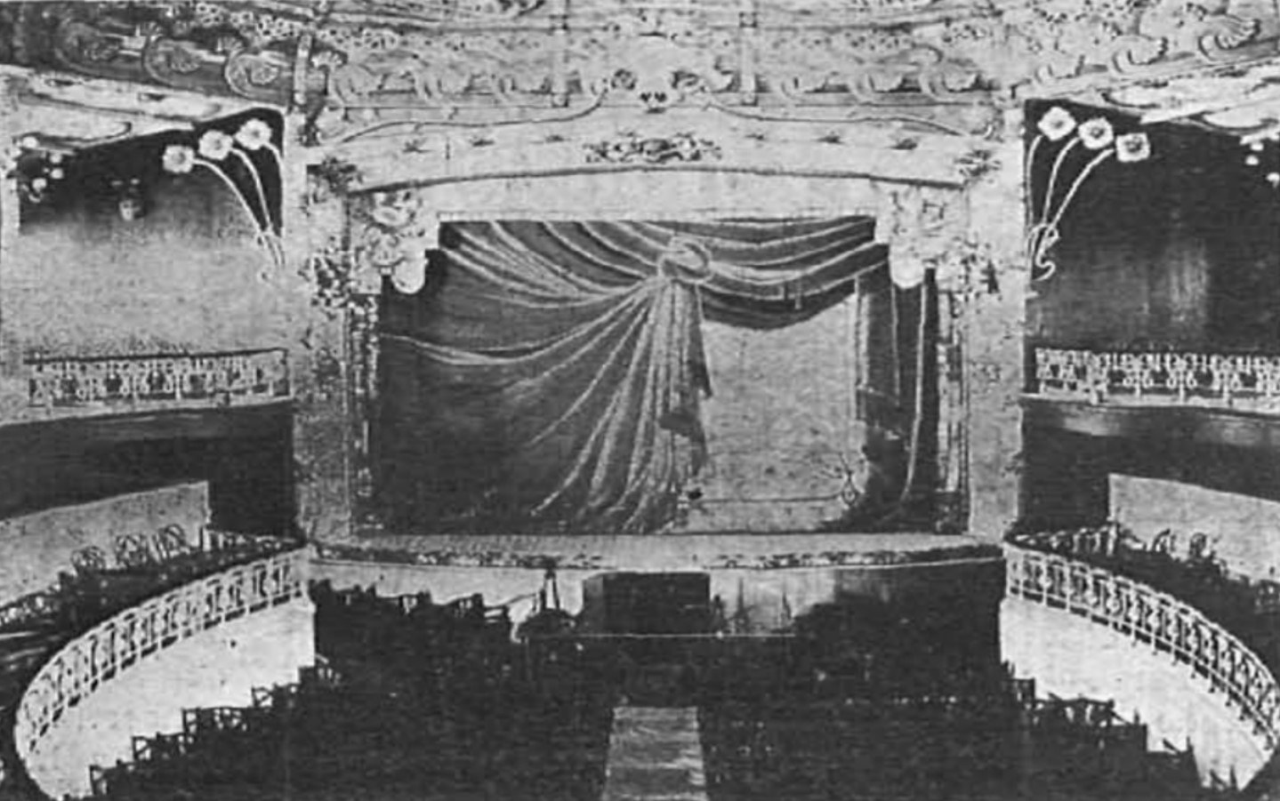 Schwarzes und weißes Foto des Auditoriums des Theatre Royal in London, das eine Bühne mit Vorhang, Geländer und ornamentierte Wandgestaltungen zeigt, mit ein paar sitzenden Menschen und Text am unteren Rand.