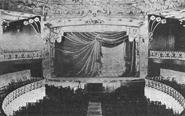 Schwarzes und weißes Foto des Auditoriums des Theatre Royal in London, das eine Bühne mit Vorhang, Geländer und ornamentierte Wandgestaltungen zeigt, mit ein paar sitzenden Menschen und Text am unteren Rand.