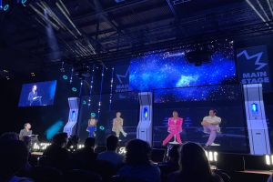 Eine Gruppe von Menschen auf einer hell erleuchteten Bühne bei einem Gaming-Event, mit einem Redner mit Mikrofon links und einem Publikum in Stühlen vor ihnen, unter einer Decke mit Beleuchtungsausrüstung.