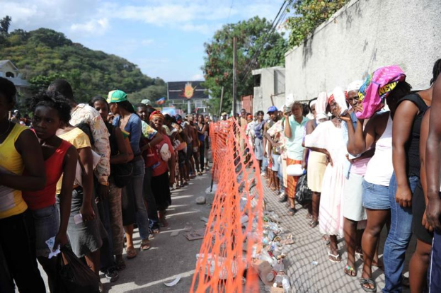 Lange Schlange von Menschen, die an einer Wahllokalwarteschlange stehen, mit einem Zaun, einer Wand, Bäumen, Gebäuden, Strommasten und einer Werbetafel im Hintergrund.