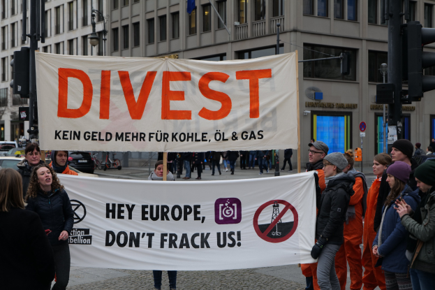 Gruppe von Menschen mit einem Banner "Divest Hey Europe, Don't Frack Us" auf einer Straße mit Gebäuden, Straßeinrichtungen, Fahrzeugen, Bäumen und einem klaren Himmel im Hintergrund.