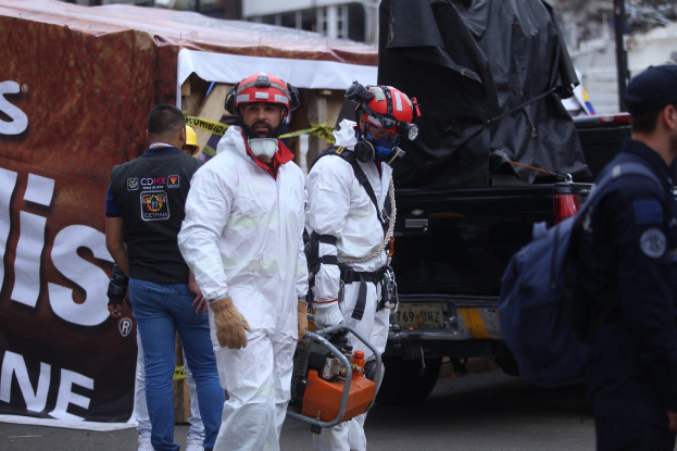 Eine Gruppe von Menschen in Schutzausrüstung, einschließlich Helmen, die eine Straße entlanggehen, mit einem Lastwagen und Gebäuden im Hintergrund, eine Person hält ein Objekt, und eine Fahne mit Text auf der linken Seite.