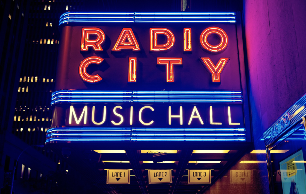 Außensicht des Radio City Music Hall in New York City bei Nacht, hell erleuchtet mit hohen Gebäuden im Hintergrund, mit einer "Radio City"-Leuchtschrift und zusätzlichen Schildern auf der rechten Seite.