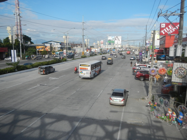 Eine belebte Stadtstraße mit verschiedenen Fahrzeugen, Strommasten, Gebäuden mit Schildern, Bäumen, Pflanzen und einem Turm unter einem bewölkten Himmel.