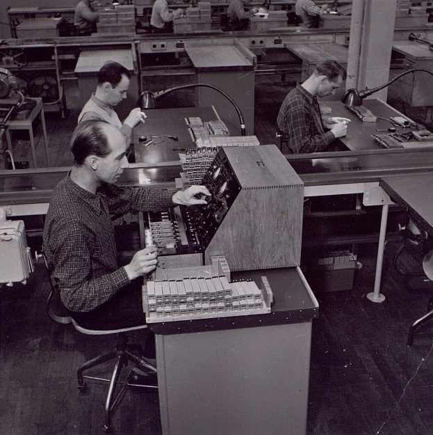 Schwarz-weißes Bild von Männern, die an Tischen arbeiten, während sie auf Computern in einer Fabrikumgebung arbeiten.