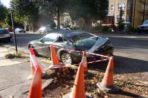 Ein verunglücktes Auto am Straßenrand, umgeben von orangefarbenen Verkehrskegeln, mit Bäumen, Gebäuden mit Fenstern und einem klaren blauen Himmel im Hintergrund.