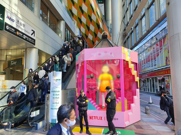 Eine Gruppe von Menschen steht vor einem Glaswand-Gebäude mit Texttafeln, einige tragen Masken, während eine Rolltreppe Menschen in der Nähe transportiert; eine pinkfarbene Box in der Mitte zeigt Schaufensterpuppen und trägt den Text "Netflix's new Netflix booth in Tokyo."
