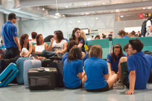 Ein belebter Flughafeninnenraum mit vielen Menschen, die auf Flüge warten, Gepäck links und Glastüren mit Richtungsschildern im Hintergrund.