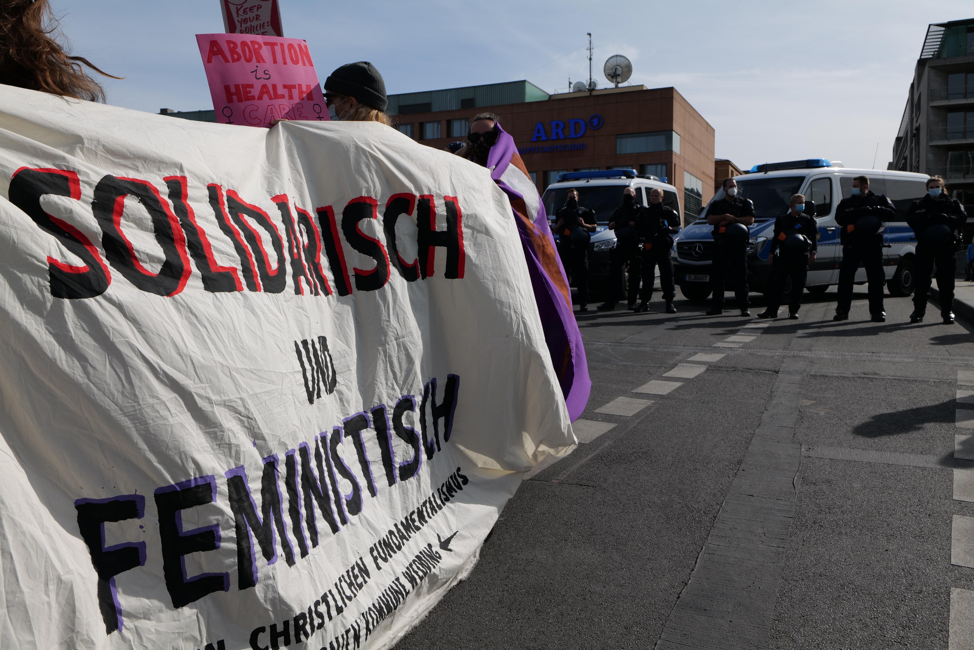 Gruppe von Menschen marschiert auf einer Straße und hält ein Banner mit der Aufschrift "Solidarität und Feminismus" mit parkenden Fahrzeugen und Gebäuden im Hintergrund bei klarem blauem Himmel.