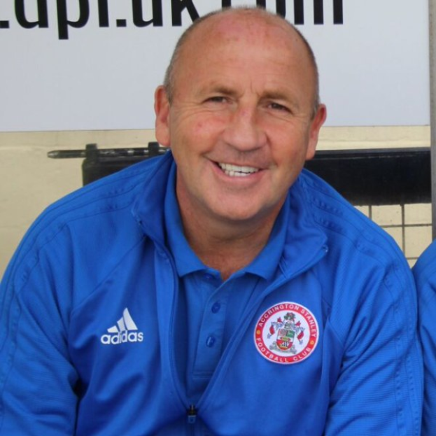 Zwei lachende Männer, einer in einer blauen Jacke, sitzen nebeneinander mit einer Wandtafel im Hintergrund, wobei der Mann in der blauen Jacke wahrscheinlich der Trainer des Accrington Stanley Football Club ist.