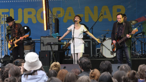 Vier Personen auf einer Bühne: Eine Frau in einem weißen Kleid singt in der Mitte, flankiert von zwei Männern, die Gitarren spielen, mit einer Menge unten und Lautsprechern an einer Wand im Hintergrund.