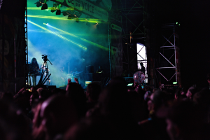 Eine Menge schaut Performern auf einer hell erleuchteten Bühne bei einem Konzert zu, mit Instrumenten, Mikrofonen und einem Banner im Dunkeln.