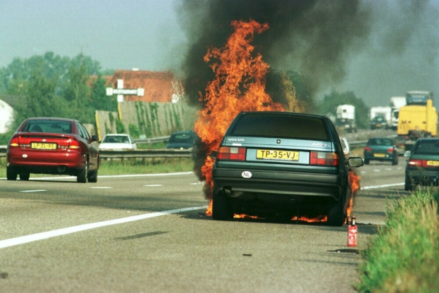 Ein Auto in Flammen auf dem Seitenstreifen, umgeben von anderen Fahrzeugen, mit Bäumen, Gebäuden und einem klaren blauen Himmel im Hintergrund und einem Feuerlöscher auf der rechten Seite des Grases.