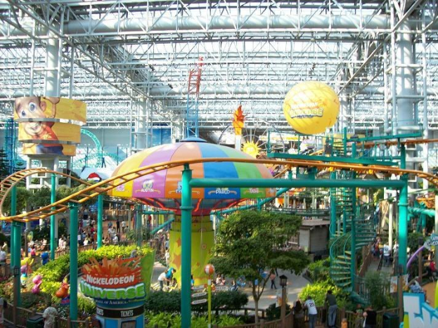 Großes Indoor-Wasserpark mit einer Achterbahn, umgeben von Menschen, Bäumen, Pflanzen und Deckenbeleuchtung mit Stangen.