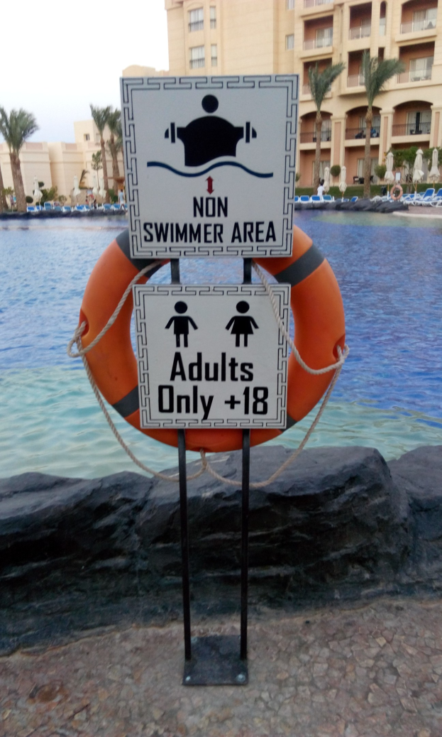 Ein Schwimmbad mit einem "Nichtschwimmer-Bereich Erwachsene Nur"-Schild im Vordergrund, umgeben von Bäumen, Gebäuden und einem klaren blauen Himmel, mit einem Rettungsring in der Mitte des Pools.