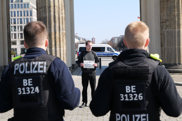 Drei Polizeibeamte stehen vor einem Gebäude, ein Mann in der Mitte hält ein Papier, flankiert von Säulen und umgeben von Fahrzeugen, Menschen, Bäumen und Gebäuden unter dem Himmel.