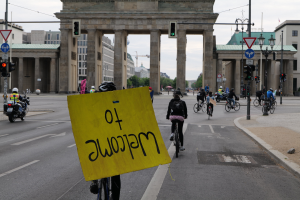 Eine Gruppe von Menschen, die auf Fahrrädern die Straße vor dem Brandenburger Tor in Berlin, Deutschland, entlangfahren, wobei eine Person ein gelbes Schild hält.