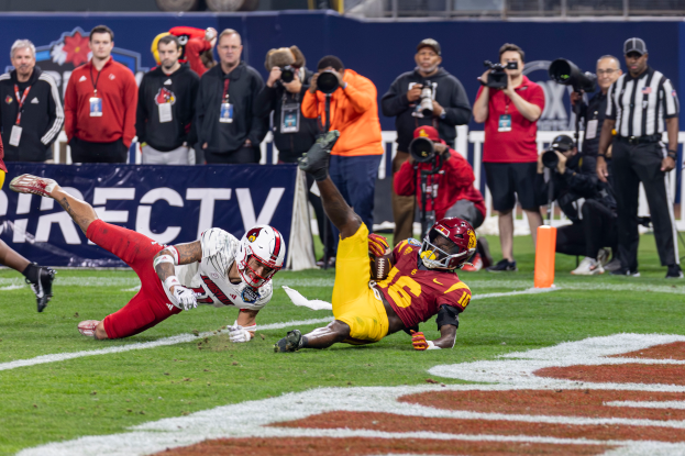 Ein Fußballspieler taucht in die Endzone für ein Touchdown ein, mit einer Gruppe von Menschen am Boden, einige halten Kameras, und ein Banner mit Text im Vordergrund, während Geländer und Pfosten im Hintergrund sichtbar sind.