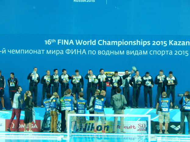 Eine Gruppe von Menschen steht am Rand eines Schwimmbeckens, einige halten Kameras, mit einem Netz im Vordergrund und einem Banner mit der Aufschrift "FINA World Championships 2015 Kazan" im Hintergrund.