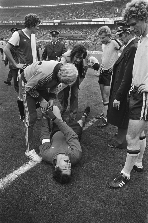 Schwarzes und wei├čes Foto eines Fussballspielers auf dem Boden umgeben von einer Gruppe von M├Ąnnern, mit einer Menge im Stadion im Hintergrund.