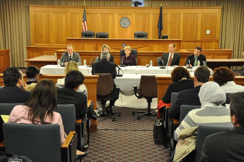 Ein Versammlungsraum mit auf Stühlen sitzenden Menschen, zwei ausländische Flaggen, ein oranger Schreibtisch mit Wasserflaschen und einem Mikrofon.