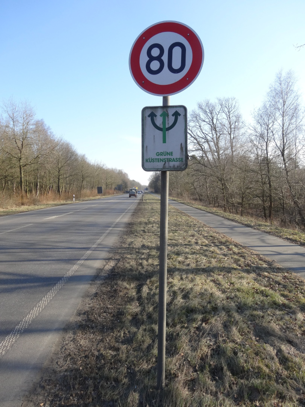 Ein Geschwindigkeitsbegrenzungsschild am Seitenrand einer Straße mit vorbeifahrenden Fahrzeugen, umgeben von Gras und Bäumen, unter einem klaren blauen Himmel.